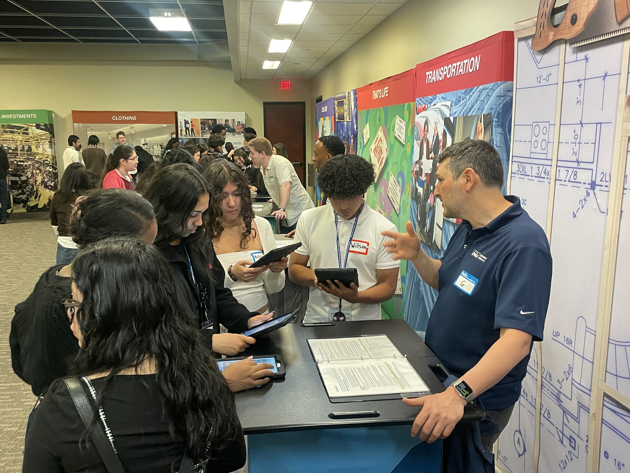 People crowd a booth at a career fair; a man in a blue polo explains to attendees beside large 'Transportation' panels and blueprint-style displays on the wall.