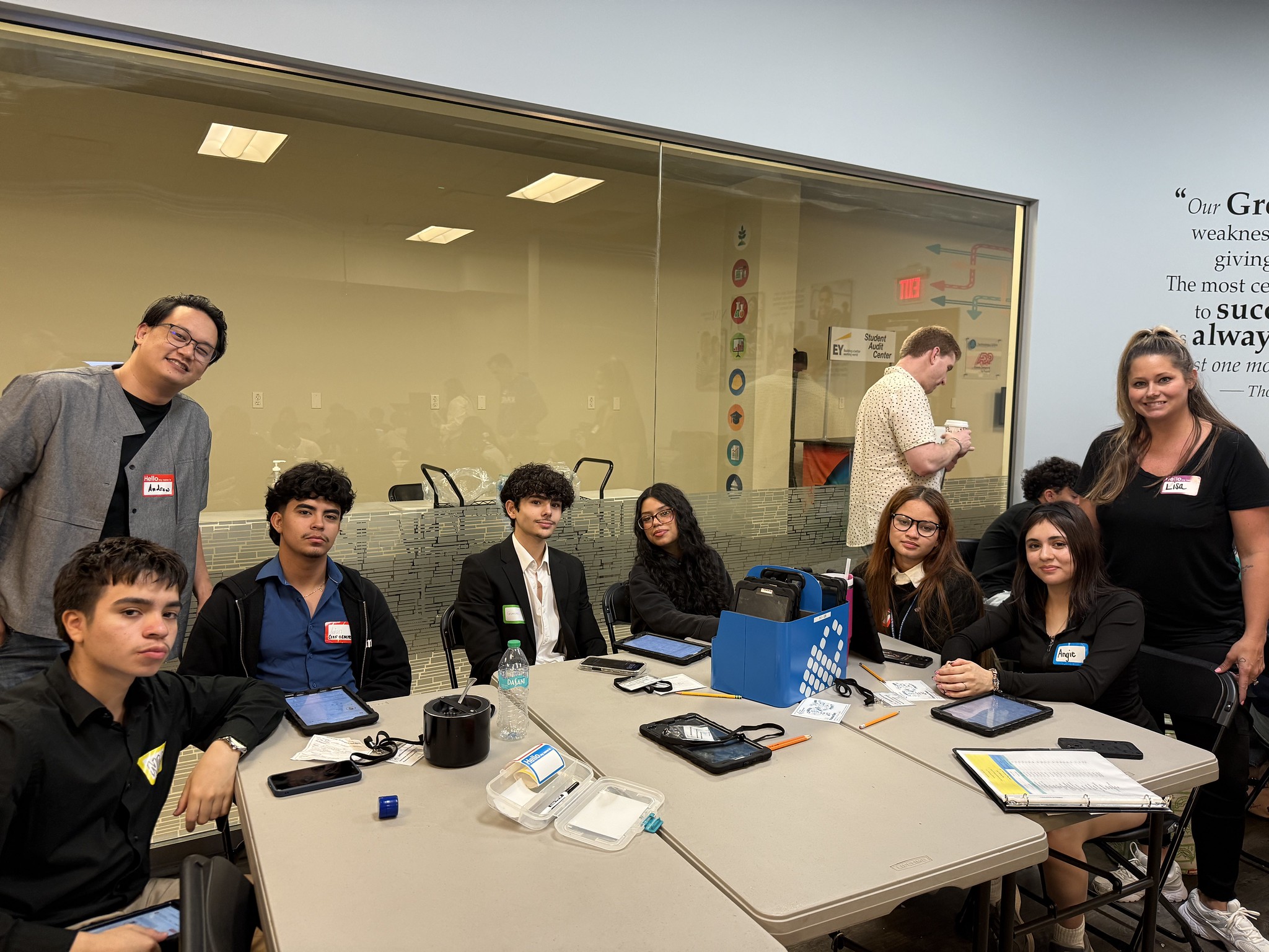 Group of students seated around a classroom table, facing the camera, with name tags and tablets on the table.