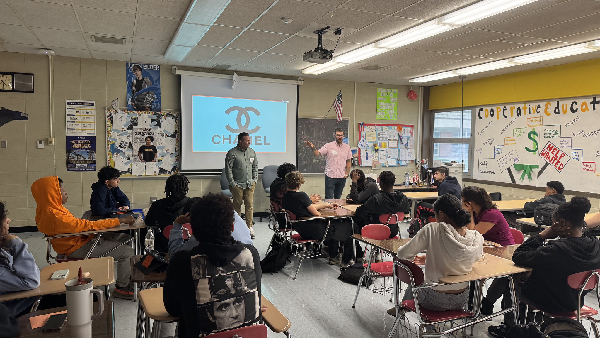 Two teachers stand at the front of a classroom presenting to students; a projector screen shows a blue Chanel logo.